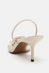 Zapato de tacón destalonado en color crudo, con cuerpo de mesh transparente con bordados florales y aplicación de abalorios brillantes. Cuenta con tira trasera elástica, tacón medio fino y acabado en punta.