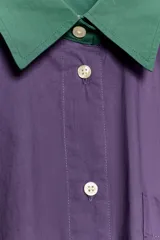 Camisa de popelín de corte oversize, color violeta con cuello y puños anchos a contraste en color verde. Presenta cierre frontal con botones, mangas largas y un bolsillo de parche en el pecho.