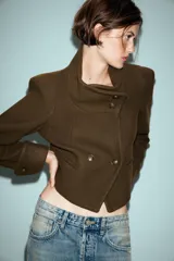 Chaqueta corta de paño color verde militar, con cuello subido, manga larga con puños abotonados y cierre frontal cruzado con botones metálicos. Presenta hombreras y bolsillos de vivo en el delantero.