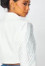 Blazer corto de corte estructurado en color blanco, con solapas clásicas y cierre frontal de un botón. Presenta un diseño decorativo con aplicaciones de pedrería brillante que recorren los hombros y las mangas en patrones curvos y geométricos.