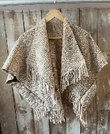 Capita tejida en 100% lana merino de color beige, con diseño abierto, cuello solapa y flecos en los bordes.