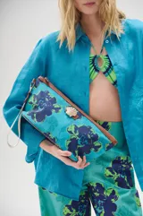 Clutch rectangular de lona estampada con flores azules y verdes sobre fondo celeste. Tiene cierre superior con cremallera y correa de mano desmontable.