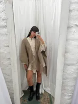 Ruana tejida a mano en lana merino color beige, con textura de punto y detalle de flecos en el ruedo. Presenta sutiles destellos brillantes integrados en el tejido.