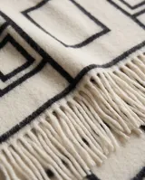 Poncho abierto color blanco marfil con diseño geométrico de rombos y cuadrados en color negro, terminado con flecos largos en el borde inferior.