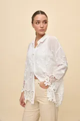 Camisa blanca de calce relajado, con cuello clásico, mangas 3/4 y cierre frontal de botones. Presenta bordados florales en relieve y detalles de crochet calado en los puños y el ruedo.
