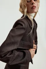 Chaqueta corta de denim oscuro con cuello solapa, cierre frontal con cremallera y bolsillos delanteros de plastrón con solapa. Presenta mangas largas con hombreras y costuras a la vista.