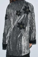 Kimono de terciopelo gris con acabado texturizado, cuello redondo y manga larga. Presenta bordados florales en hilo negro y metalizado, bolsillos delanteros y cierre frontal.