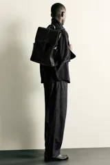 Mochila de diseño minimalista en color negro, confeccionada en material sintético con acabado liso. Presenta una solapa frontal con cierre de hebilla decorativa y sistema de imán, compartimento principal con cremallera, bolsillos laterales externos, asa superior de mano y correas de hombro ajustables y acolchadas.