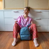 Mochila de tamaño mediano en color amarillo, confeccionada en símil cuero. Presenta un compartimento principal con cierre metálico, un bolsillo frontal con cierre y broche, y bolsillos laterales. Incluye correas regulables para la espalda, asa de mano y una correa adicional para llevar al hombro.