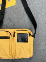 Cartera bandolera amarilla de nylon con correa negra ajustable. Tiene dos bolsillos frontales con cierre y un parche negro con el logo de la marca.
