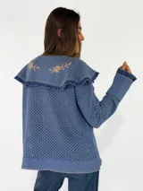 Saco tejido de punto color azul con diseño calado, cuello grande con bordados florales en tono crudo, volados decorativos y cierre frontal con un botón.