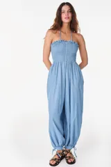 Mono largo de denim color celeste, sin mangas, con escote nido de abeja elástico y volado superior, tirantes finos para atar al cuello, perneras amplias de corte abombado con puños ajustables mediante lazos y bolsillos laterales.