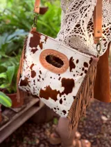 Bandolera de cuero con estampado animal print de vaca en tonos marrón y blanco, con flecos de cuero marrón en los laterales. Tiene correa larga de cuero marrón regulable y asa de mano con forma ovalada.