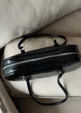 Cartera de hombro tipo baguette en color negro con acabado brillante efecto cuero arrugado. Presenta una estructura rectangular con bordes redondeados, cierre superior y doble asa fija.