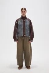 Campera estilo bomber con diseño color block, combinando paneles de denim lavado con mangas y detalles en cuero sintético color marrón oscuro. Presenta cuello alto con tira ajustable, cierre frontal con botones a presión y bolsillos frontales tipo parche.