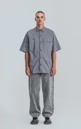 Camisa de manga corta en color gris, confeccionada en popelina de algodón. Presenta un corte oversize, cuello clásico, cierre frontal con botones y dos bolsillos delanteros, uno de ellos con solapa.