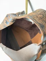 Cartera tipo hobo de cuero genuino con diseño bicolor que combina paneles de cuero liso marrón con paneles de cuero texturizado con efecto desgastado en tonos negro y marrón. Incluye un sobre extraíble a juego con cierre y etiqueta de la marca.