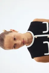 Chaleco corto negro en tejido bouclé, con cuello redondo, cierre frontal de botones dorados y bolsillos frontales decorativos ribeteados en blanco.