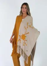 Ruana de tejido de punto bicolor, combinando mostaza y beige, con diseño abierto, bordado floral en relieve y flecos en el ruedo.