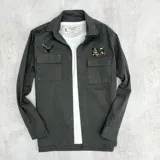 Sobrecamisa gris de gabardina con cuello clásico, cierre frontal con botones, dos bolsillos de parche con solapa en el pecho, mangas largas con puños abotonados y parches decorativos.