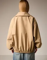 Chaqueta color beige de estilo utilitario con cuello alto y cierre frontal parcial. Presenta mangas amplias con puños elásticos y dos bolsillos frontales tipo cargo con solapa.