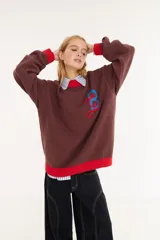 Sweater de punto color amarillo con silueta oversize y estilo universitario vintage. Presenta cuello redondo, puños y ruedo en tejido acanalado de color gris azulado, y un diseño gráfico bordado en el frente.