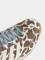 Championes Adidas modelo SL 72 OG con diseño de perfil bajo. Presentan una parte superior de piel con acabado de pelo de poni con estampado animal print de manchas marrones y blancas. Incluyen las icónicas tres tiras laterales en color crema, cordones celestes y suela de goma con entresuela de EVA.