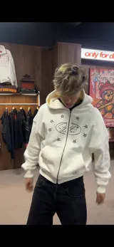 Buzo con cierre frontal, color crema, con diseño estampado de estrellas y logo en el pecho. Cuenta con capucha, bolsillos tipo canguro y tirador de cierre en forma de estrella.
