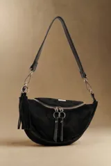 Cartera tipo bandolera de diseño semicircular en color negro, confeccionada en material sintético con textura de gamuza. Cuenta con cierre frontal con doble tirador metálico circular, borla decorativa lateral y correa ajustable con herrajes plateados.