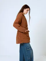 Campera de pana color marrón, con diseño acolchado, cuello camisero, cierre frontal oculto y cinturón ajustable con hebilla a tono. Presenta bolsillos frontales con solapa y mangas largas de corte holgado.