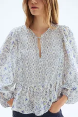Blusa en mezcla de viscosa con peplum acampanado, cuello redondo, escote en V y tiras finas con cuentas decorativas. Presenta mangas globo largas con elástico en los puños y un estampado en tonos blancos y azules con motivos florales y geométricos.