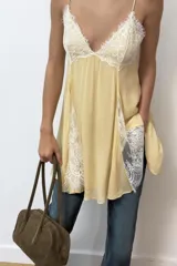 Vestido corto amarillo con escote en pico y tirantes finos ajustables. Presenta detalles de encaje blanco en el escote y en los laterales de la falda. Tiene forro interior y cierre en la espalda con cremallera y botones forrados.