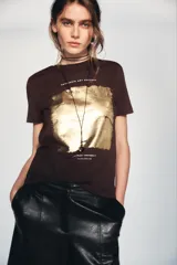 Camiseta de algodón color marrón oscuro, de corte regular, cuello redondo y manga corta. Presenta una estampación metalizada dorada en el parte frontal con texto "REYKJAVIK ART GALLERY" y "ABSTRACT MOVEMENTS Reykjavik, Iceland, 1975".