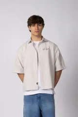 Camisa de manga corta de popelina con diseño a rayas verticales en tonos beige y blanco, corte boxy, cuello camisero y cierre frontal de botones. Cuenta con bordado sutil en el pecho y etiqueta institucional en el ruedo.