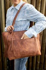 Cartera tipo tote de cuero marrón con costuras artesanales visibles, bolsillo frontal con el logo grabado, asas de hombro y detalles de flecos colgantes.