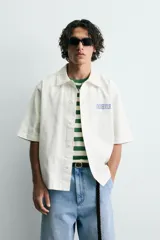Camisa de corte relajado color verde oliva, con cuello solapa, manga corta y cierre frontal de botones. Presenta un estampado con el texto 'ASTRAL Drift' en el frente.