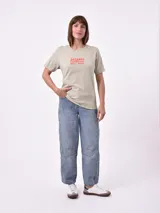 Remera de manga corta color beige claro con cuello redondo. Presenta un estampado gráfico central con un plato de pasta rodeado por un borde azul y blanco, sobre un fondo de franjas verticales rojas y blancas. El estampado incluye texto en cursiva en tonos turquesa y rojo.