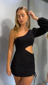 Vestido corto de lurex negro con diseño asimétrico de un solo hombro y manga larga abullonada. Presenta un recorte lateral en la cintura y detalle de fruncido con cordón ajustable en el ruedo.