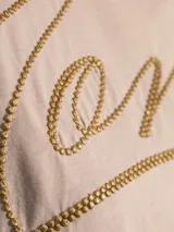 Remera de algodón color rosa pálido, con cuello redondo y mangas cortas. Presenta el logo de la marca bordado en el frente con un diseño de tipografía cursiva en hilo dorado.