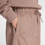 Pantalón deportivo New Balance modelo Woven Pant, color marrón topo, de tejido ligero y corte ancho. Presenta cintura elástica con cordón ajustable y un pequeño logo New Balance bordado en la pierna izquierda.