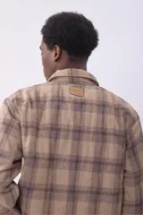 Campera tipo sobrecamisa de franela con diseño a cuadros en tonos beige y marrón. Presenta cuello clásico, cierre frontal con botones, dos bolsillos frontales con solapa y botones en el pecho, y mangas largas con puños abotonados.