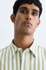 Camisa de popelina de algodón con cuello inglés, tapeta clásica, bolsillo superior abierto y canesú con pliegue en la espalda. Cuenta con mangas largas, puños ajustables con botón y bajo ligeramente redondeado. Presenta un diseño de rayas verticales en tonos verde y blanco.