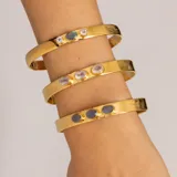 Brazalete rígido dorado con tres piedras incrustadas: una labradorita ovalada en el centro y dos piedras de la luna redondas a los lados.