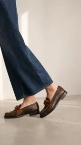 Mocasín de cuero vacuno con textura gastada en tono marrón oscuro. Presenta un detalle de banda frontal en pelo sintético al tono y costuras reforzadas en el empeine.