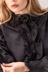 Camisa negra de corte entallado con volados decorativos en el cuello, la botonadura frontal, los hombros y las mangas. Presenta mangas largas con puños ajustados y un ruedo con volado inferior.