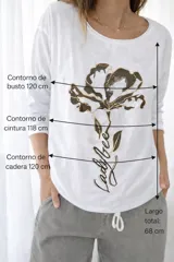 Remera blanca de línea italiana, confeccionada en algodón y viscosa, con calce holgado y cuello redondo. Presenta un estampado frontal de una flor en tonos oscuros con detalles dorados y la frase "Ladybee". Incluye tiras regulables en la parte inferior para ajustar el largo.