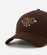 Gorra de sarga de algodón color marrón oscuro con visera curva y ajuste trasero. Presenta un bordado frontal con las letras 'NYC' en estilo universitario con detalles en rojo y blanco, flanqueadas por los números '19' y '86' y tres estrellas blancas debajo.