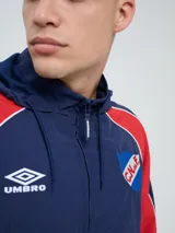 Campera deportiva Umbro del Club Nacional de Football, modelo Venti, para hombre. Diseño en color blanco con mangas azules y detalles de líneas rojas. Cuenta con capucha con cordón ajustable, cierre frontal y bolsillos. Presenta el logo de Umbro y el escudo del club bordados en el pecho.