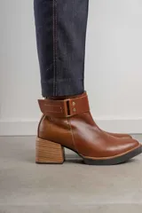 Bota corta de cuero negro con acabado combinado mate y charol. Presenta una tira decorativa con tachas metálicas y hebilla en el tobillo, tacón cuadrado robusto y suela con plataforma.