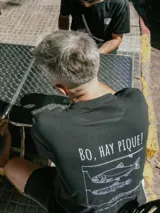 Remera negra de algodón con estampado gráfico en la espalda que incluye el texto 'Bo, hay pique!' y dibujos de peces con una caña de pescar.